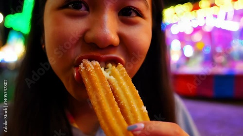 A young Hispanic woman takes a bite of churros at a vibrant carnival, surrounded by colorful lights and festive decorations