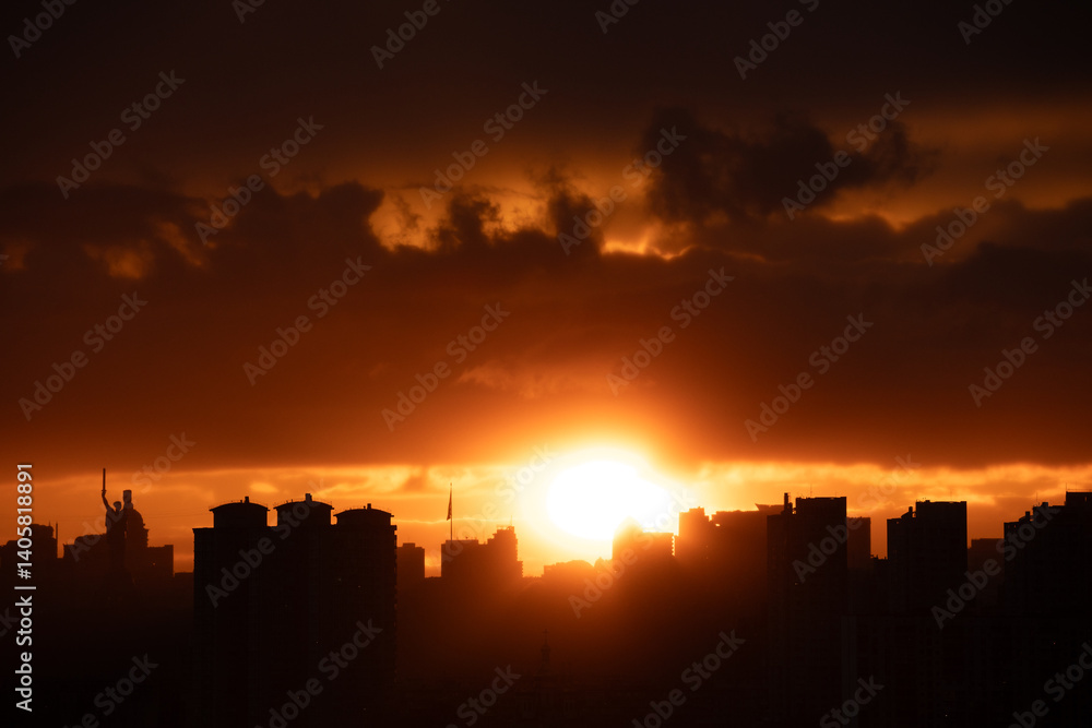 Naklejka premium A dramatic sunset over the silhouetted skyline of Kyiv, featuring both the Motherland Monument and a tall flagpole with the Ukrainian flag waving prominently.