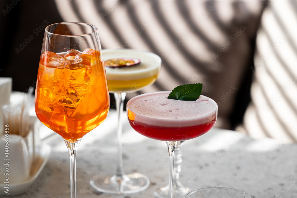 Three beautifully crafted cocktails are displayed on a marble surface, featuring vibrant colors. The drinks include an orange spritz, a creamy yellow blend, and a red beverage garnished with a leaf.
