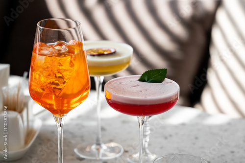 Obraz na płótnie Three beautifully crafted cocktails are displayed on a marble surface, featuring vibrant colors. The drinks include an orange spritz, a creamy yellow blend, and a red beverage garnished with a leaf. w salonie