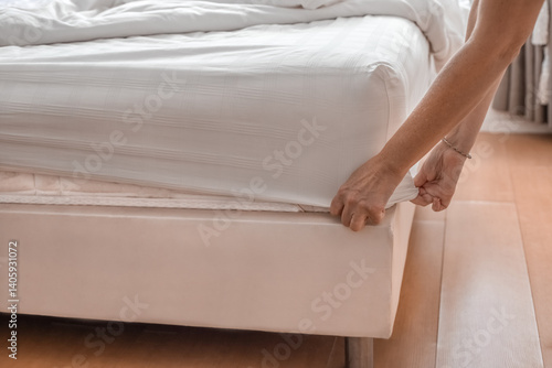 hands of woman making bed, tucking in fitted sheet and bed linen in lighted room with large window. Hotel room cleaning