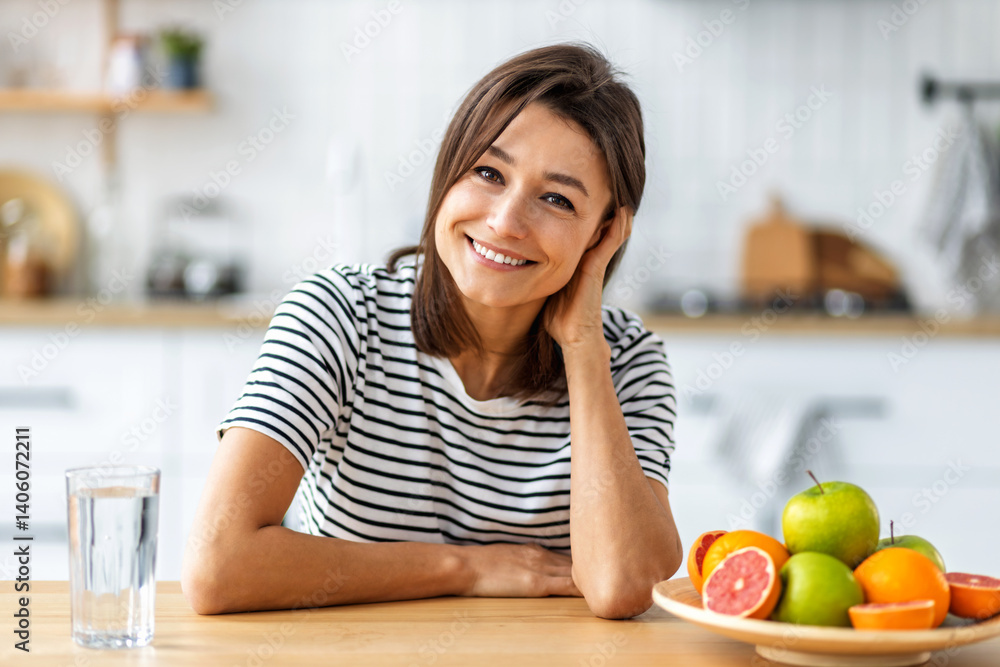 Cheerful woman with glass of water and fresh fruits smiling at camera while sitting in modern kitchen promoting healthy lifestyle and balanced nutrition