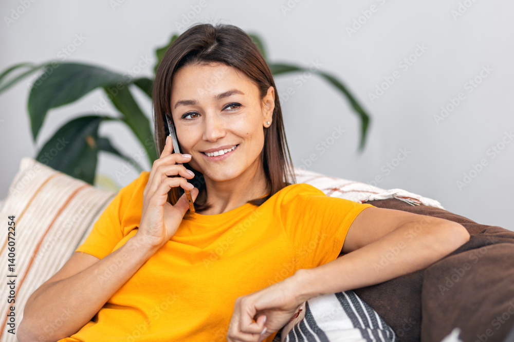 Cheerful young woman relaxing on sofa at home talking on mobile phone smiling and enjoying friendly conversation indoors