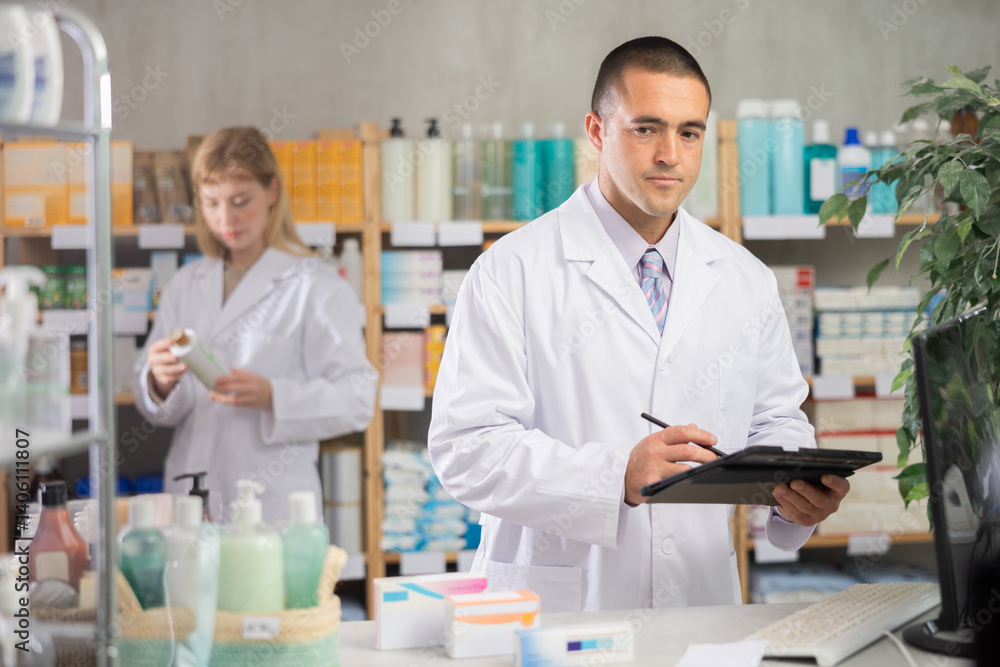 Attentive young male pharmacist restocking with tablet while female pharmacist working in background in drugstore Attentive young male pharmacist restocking with tablet while female pharmacist working in background in drugstore