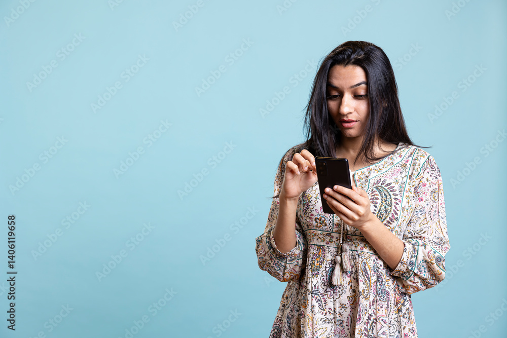 Indian woman happily scrolling on phone to check social media feed, amazed by something. Young relaxed person browsing the internet websites, texting friends against studio background.