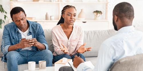 Displeased black woman blaming husband at psychologist consultation, complaining about his smartphone addiction, indifferent man spending time with phone next to annoyed wife, selective focus