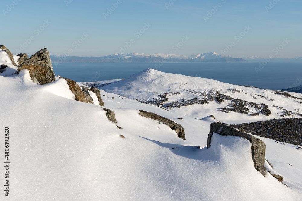 Beautiful mountain landscape. View from the snowy mountainside to the sea coast. In the foreground are rocks and white snow. The Sea of Okhotsk is in the distance. Cold weather. Magadan region, Russia