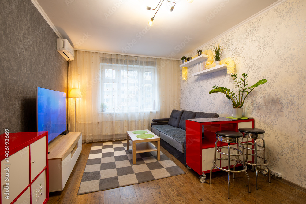 Apartment interior. Living room with furniture: dark gray sofa, red chest of drawers, metal chairs, wooden coffee table, shelves with indoor plants, TV. The window is covered with a tulle curtain.