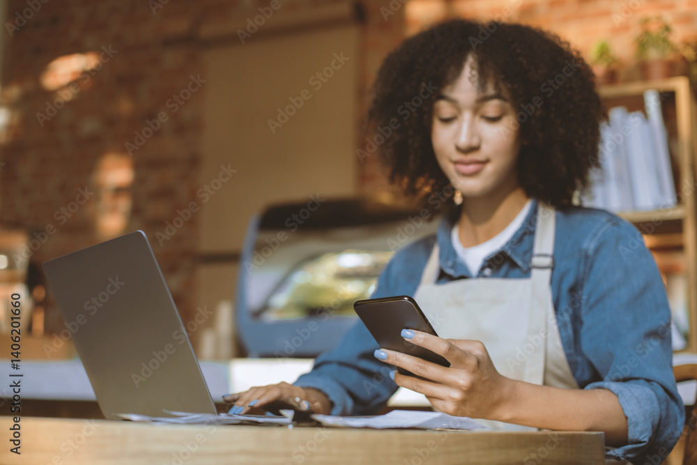 Social distancing, online order takeaway and work during coronavirus pandemic. Young african american lady owner in apron sits at table and works with laptop and phone in interior of modern cafe Social distancing, online order takeaway and work during coronavirus pandemic. Young african american lady owner in apron sits at table and works with laptop and phone in interior of modern cafe