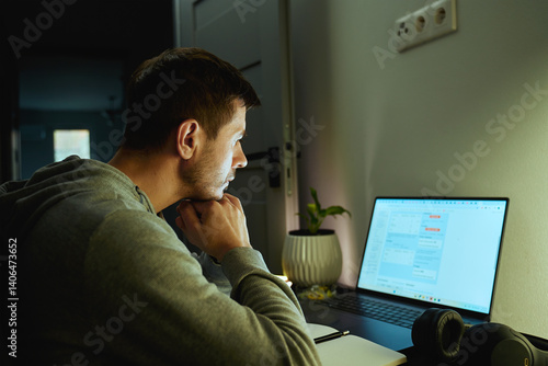 Concentrated freelancer working late at home office. Focused man in hoodie writes notes while working on laptop. Concept of night work
