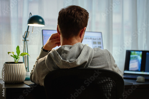 Obraz na płótnie Man in gray hoodie working at computer desk with dual monitors and lamp in background. Freelancer working remotely at home office. Concept of remote work and productivity w salonie
