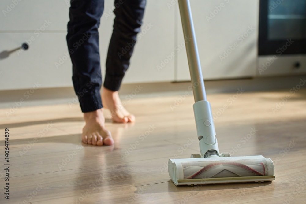 Barefoot man cleaning wooden floor in kitchen with vacuum. Person vacuuming with cordless vacuum cleaner. Daily routine and household chores