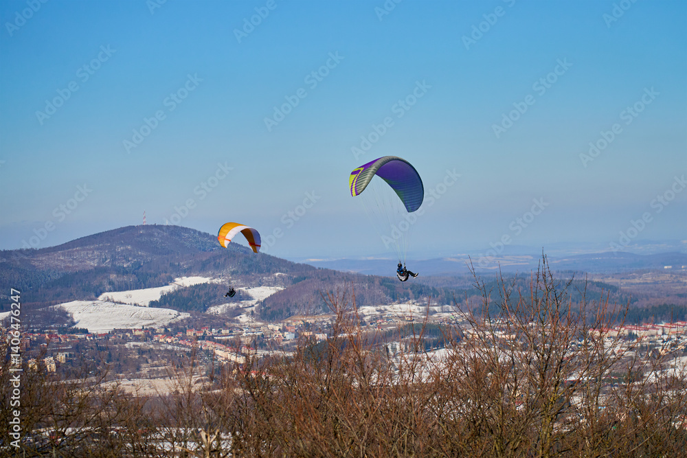 Two paragliders fly above snowy landscape with mountains and town on clear day. Concept of freedom, adventure, and outdoor sports