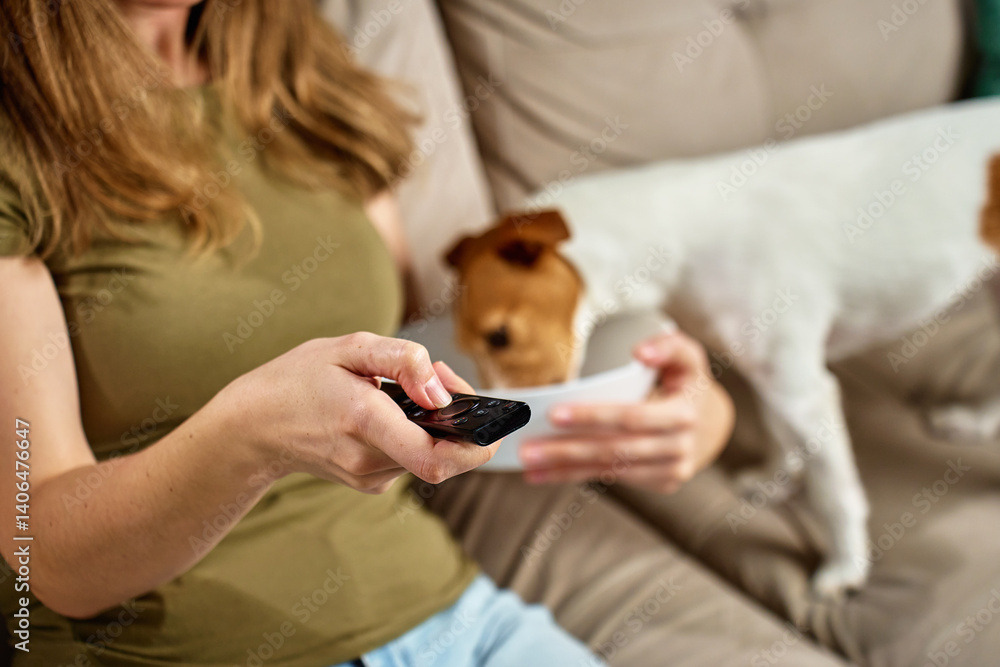 Woman relaxing on sofa with remote control while dog steals food. Pet dog eat food from bowl while owner focuses on TV. Home life and pet behavior concept