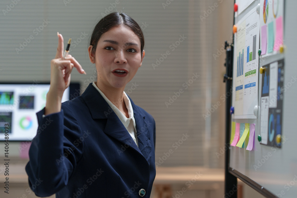 Young businesswoman explaining business strategy using whiteboard and sticky notes during a corporate meeting with colleagues working on computers in the background Young businesswoman explaining business strategy using whiteboard and sticky notes during a corporate meeting with colleagues working on computers in the background