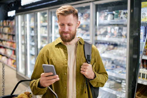 Obraz na plátně Man holding phone and shopping basket while checking grocery list in supermarket