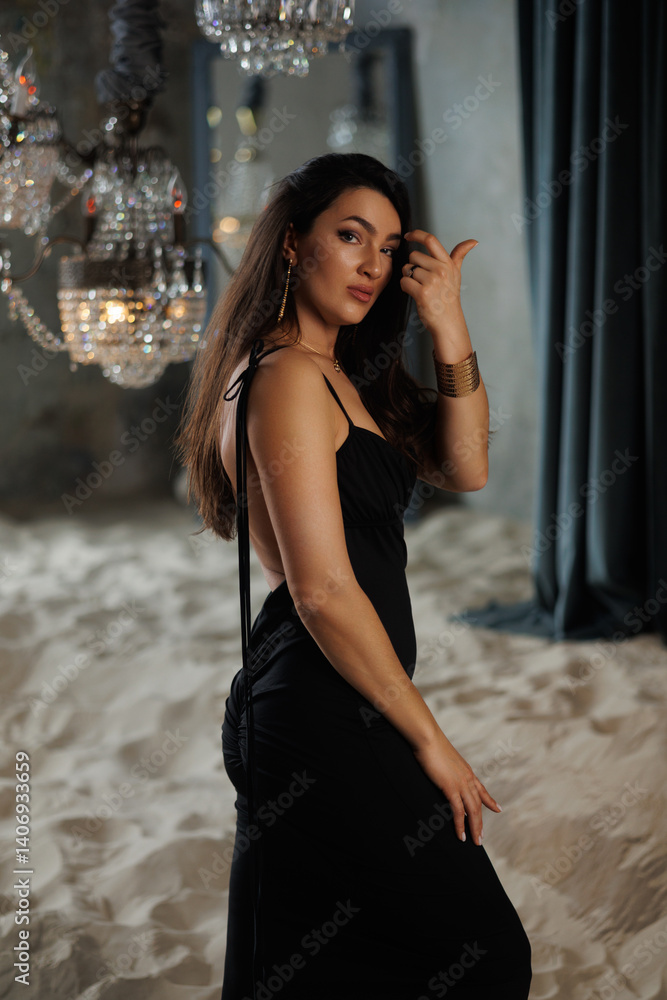 Obraz premium beautiful brunette with thick hair in a long black dress in a studio with sand and chic chandeliers. Gray walls and floor-to-ceiling windows. Elegant woman of 30 years old, athletic figure. Portrait
