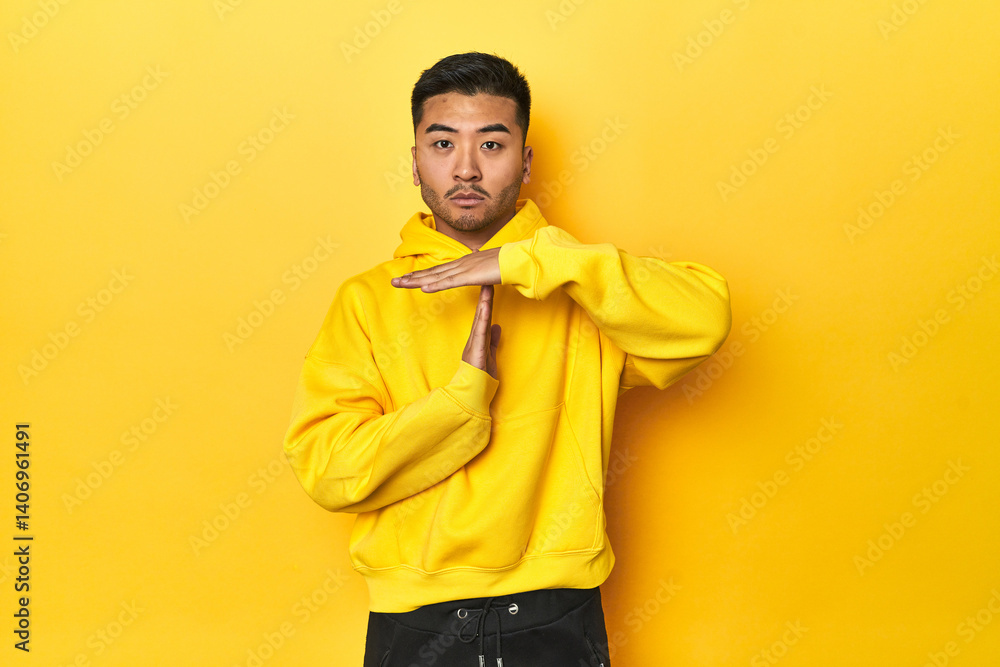 Fototapeta premium Bold Asian man in yellow hoodie on yellow studio showing a timeout gesture.