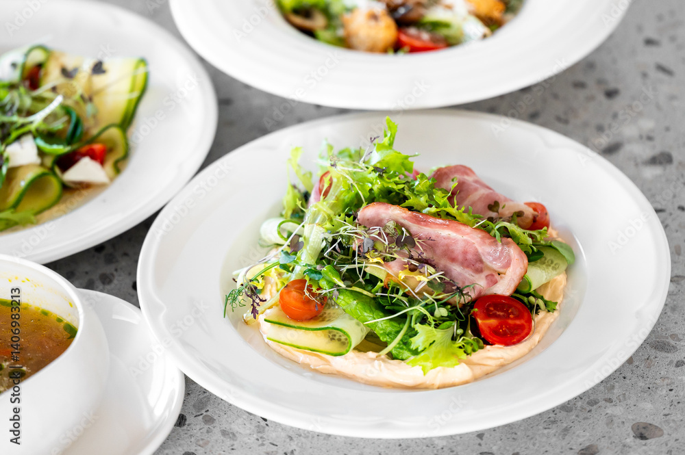 A vibrant salad featuring fresh greens, cherry tomatoes, and slices of ham on a white plate. In the background, other gourmet dishes are displayed, highlighting a beautiful dining setup. A vibrant salad featuring fresh greens, cherry tomatoes, and slices of ham on a white plate. In the background, other gourmet dishes are displayed, highlighting a beautiful dining setup.