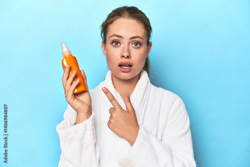 Fototapeta premium Blonde in bathrobe with sunscreen cream in studio pointing to the side