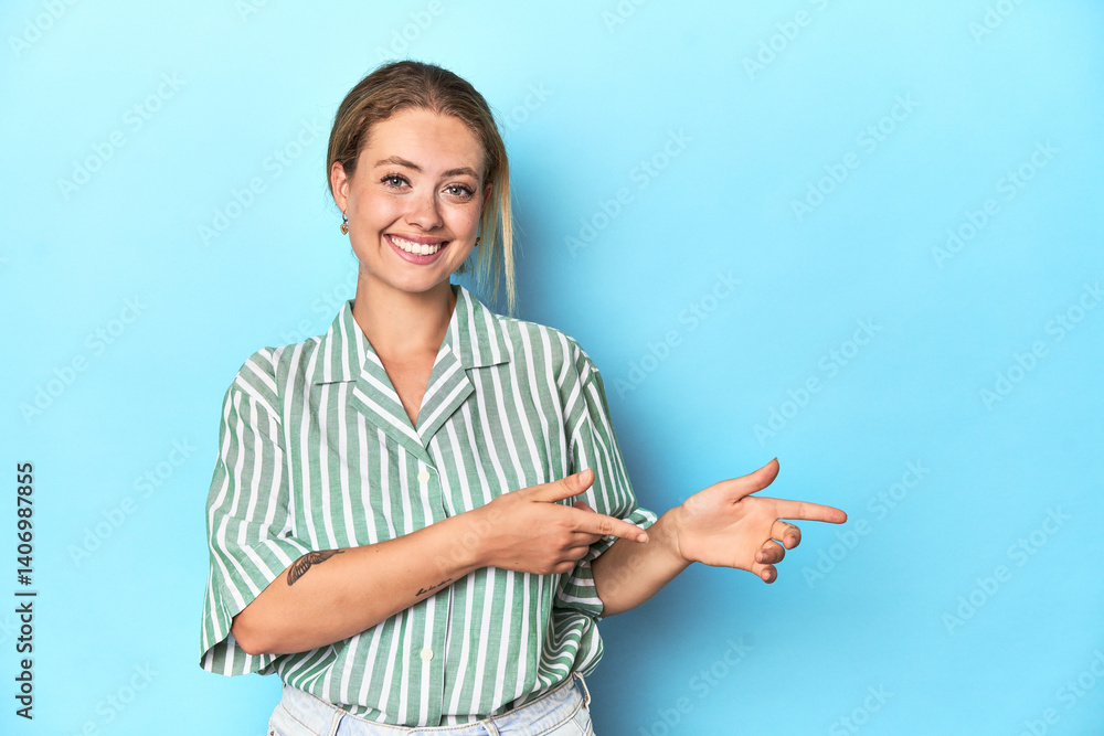 Blonde young woman in green striped shirt on blue background excited pointing with forefingers away. Blonde young woman in green striped shirt on blue background excited pointing with forefingers away.