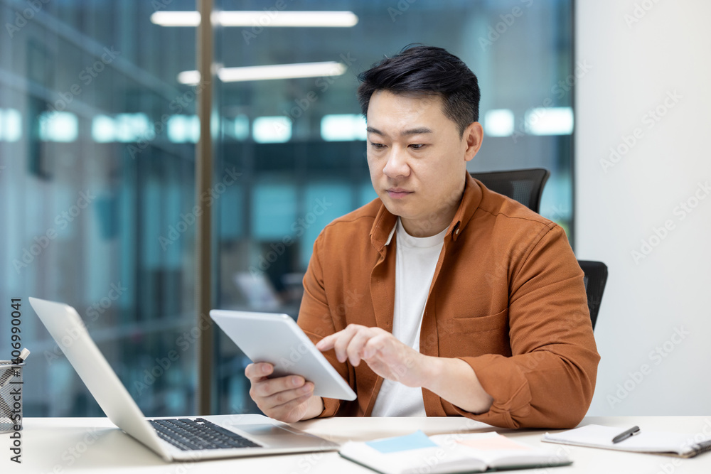 An asian businessman is seen at his desk, using a tablet. He is working on a laptop as well, focused on his tasks. An asian businessman is seen at his desk, using a tablet. He is working on a laptop as well, focused on his tasks.