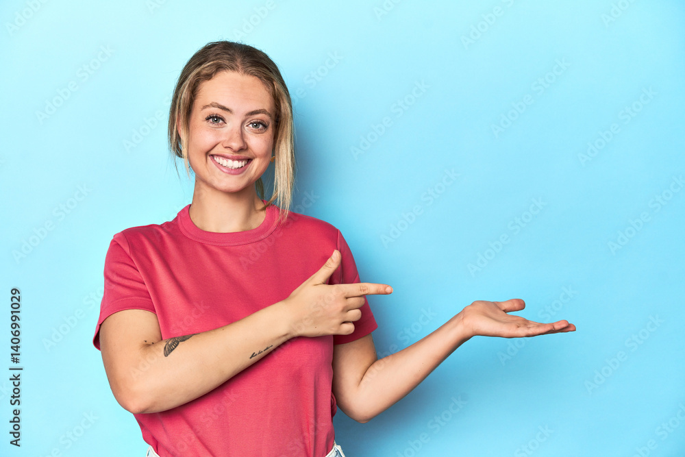 Blonde young woman in red t-shirt on blue background excited holding a copy space on palm.