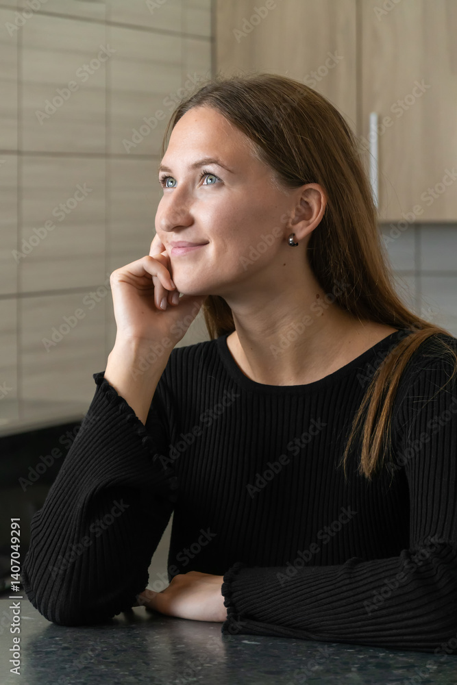 Young woman sits thoughtfully in her kitchen, resting her chin on her hand and gazing upward with a gentle smile, contemplating future plans and aspirations in a moment of peaceful daydreaming