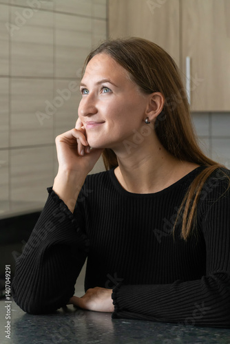 Obraz na płótnie Young woman sits thoughtfully in her kitchen, resting her chin on her hand and gazing upward with a gentle smile, contemplating future plans and aspirations in a moment of peaceful daydreaming w salonie