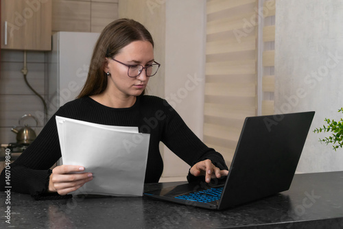 Obraz na płótnie Young woman wearing glasses, sitting at a kitchen table, working on a laptop while reviewing paperwork, managing home finances, and balancing remote work with daily life w salonie
