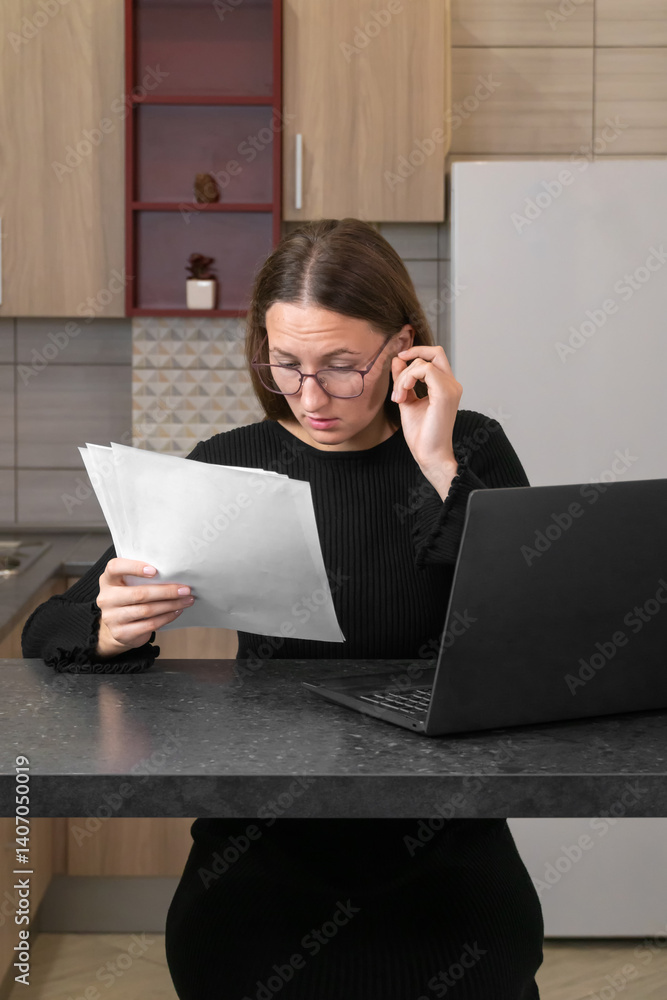 Young woman wearing glasses and working from home, holding documents and entering data into laptop, focused on her task in a bright home office setting. Vertical photo