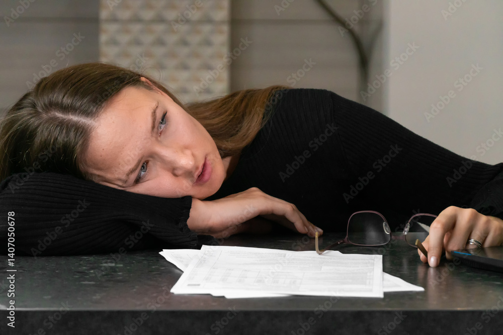 Young exhausted accountant is resting her head on the desk after working with financial documents and using laptop, showing fatigue and stress