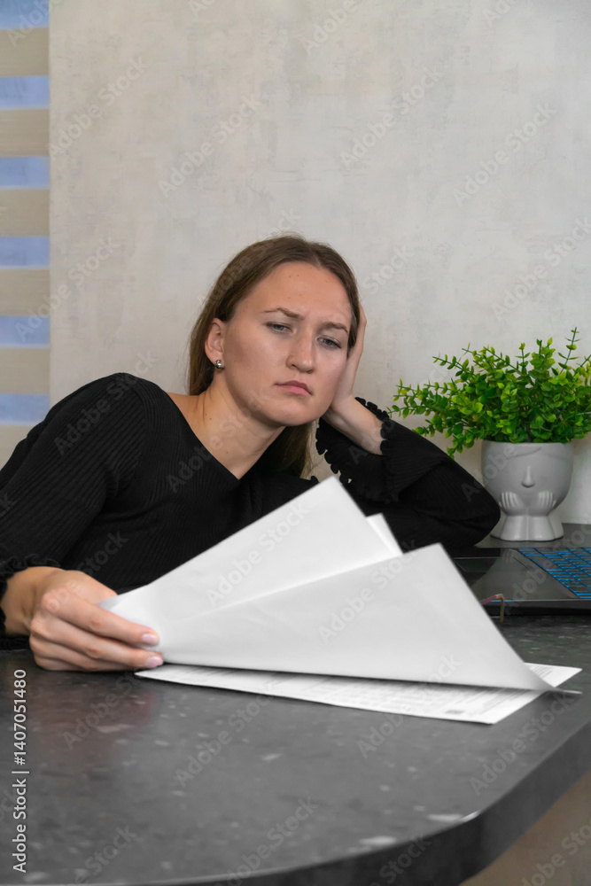 Young businesswoman resting her head on her hand, feeling tired while reading through important documents at her home office desk, with a laptop open nearby. Vertical photo