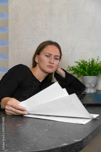 Obraz na płótnie Young businesswoman resting her head on her hand, feeling tired while reading through important documents at her home office desk, with a laptop open nearby. Vertical photo w salonie