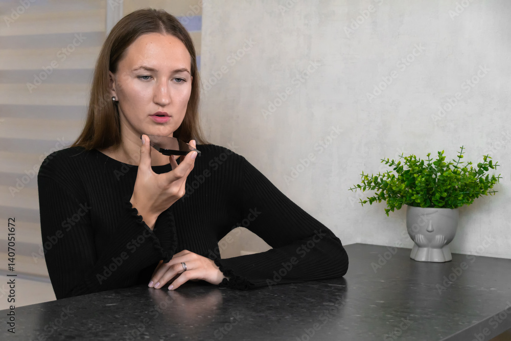 Young woman sitting comfortably at home, using her smartphone to record an audio message while talking on speakerphone, enjoying a relaxed moment in a bright kitchen