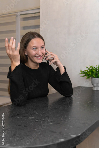 Obraz na płótnie Happy young woman is sitting at a table, talking on her phone and gesturing with her hand, enjoying a pleasant conversation in the comfort of her home. Vertical photo w salonie
