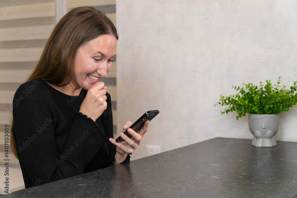 Young woman sitting at a kitchen table, laughing joyfully while reading a funny message on her smartphone, with a decorative plant nearby, enjoying a relaxed moment at home
