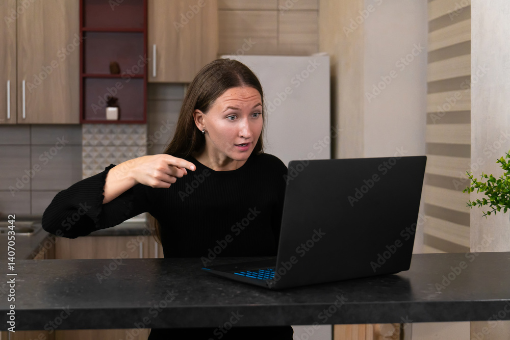 Young woman sitting at the kitchen counter, looking shocked while pointing at the laptop screen. Emotions of surprise and concern reflecting her remote work experience