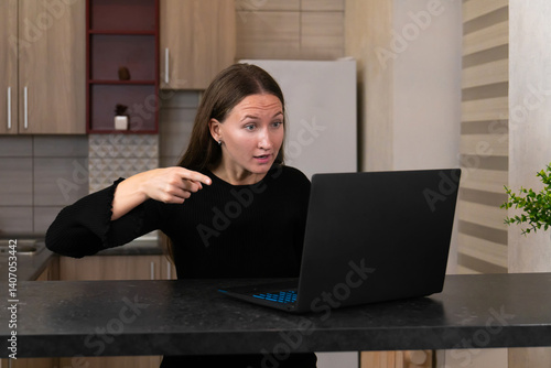 Obraz na płótnie Young woman sitting at the kitchen counter, looking shocked while pointing at the laptop screen. Emotions of surprise and concern reflecting her remote work experience w salonie