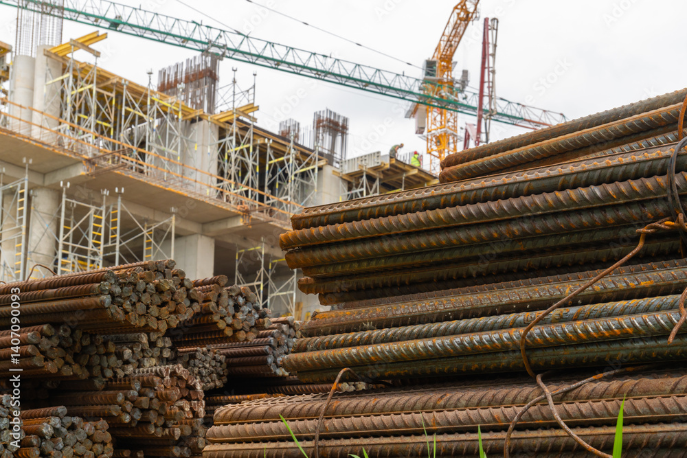 Steel rods stacked together at a construction site, with construction workers and a crane visible working on the building in the background