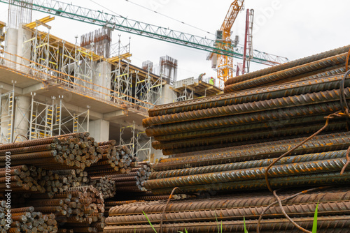 Obraz na płótnie Steel rods stacked together at a construction site, with construction workers and a crane visible working on the building in the background w salonie