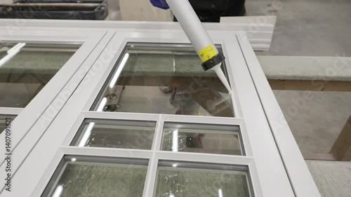 A worker in gloves carefully applies sealant to a window frame using a caulking gun, ensuring a clean, tight bond between the glass and the white frame