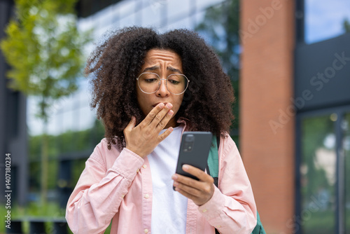 A young woman in glasses is surprised by something on her phone, covering her mouth with her hand outdoors. Modern building in background.