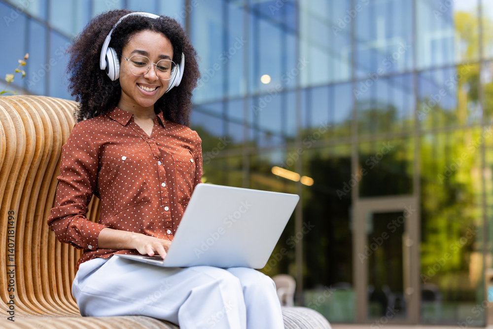A young woman with headphones smiles while working on a laptop outdoors, near a modern building. She is dressed professionally in a polka dot shirt and glasses.