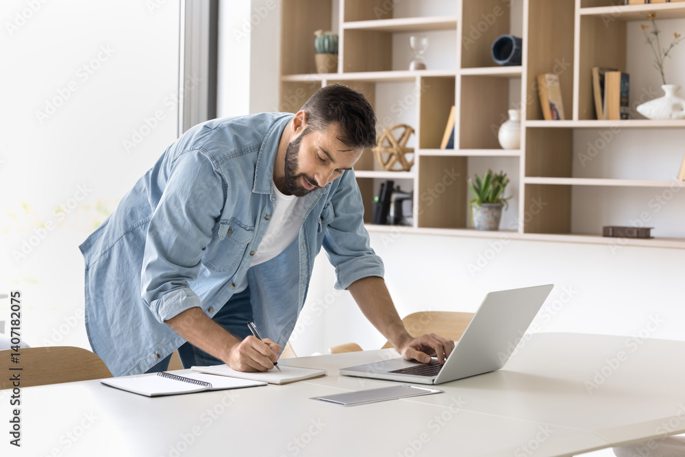 Home office workflow. Focused millennial man studying or working on distance stand by workplace table browse internet for information write notes in paper copybook managing tasks planning work process Home office workflow. Focused millennial man studying or working on distance stand by workplace table browse internet for information write notes in paper copybook managing tasks planning work process