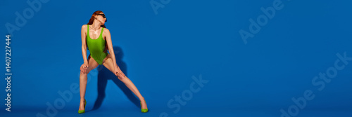 Flyer. Tall slim girl wearing in bright green swimsuit doing stylish split pose in heels against blue backdrop, expressing boldness and power. Concept of summer, vacation, holidays, fashion and style.