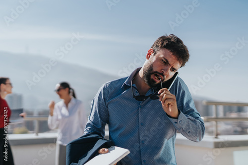 Obraz na płótnie A focused businessman multitasks outdoors, holding a phone and chewing on his glasses. Colleagues engage in the background, depicting a busy professional environment. w salonie