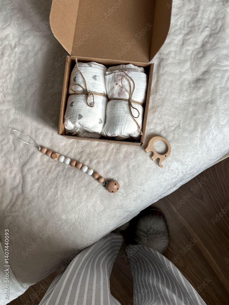 A girl is photographing wrapped delicate muslin diapers in a box, next to beads, a wooden elephant teether is lying on the bed. View from above