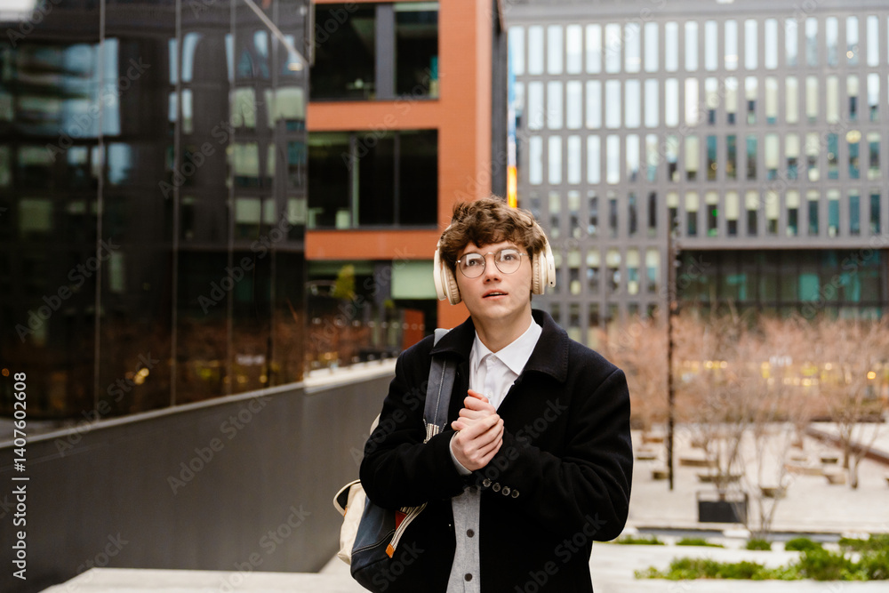 Outdoors in a city, near modern buildings, a White man in his 20s with curly brown hair and glasses, wearing a white shirt, grey cardigan, black coat, and white headphones, stands looking slightly up.