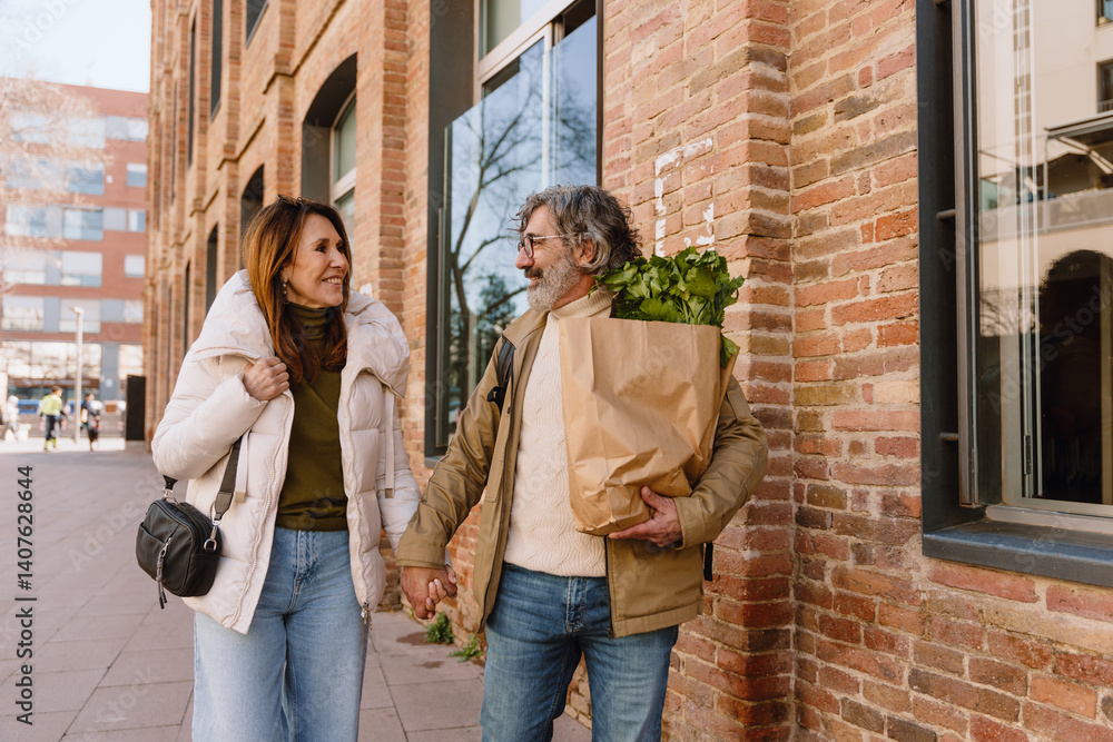 In a city near a brick building, a White couple in their 50s walks holding hands and looking at each other, a man wears a beige jacket and carries a bag of celery, and a woman wears a white jacket. In a city near a brick building, a White couple in their 50s walks holding hands and looking at each other, a man wears a beige jacket and carries a bag of celery, and a woman wears a white jacket.